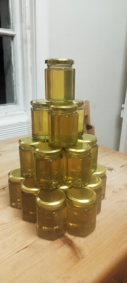 A pyramid of small hexagonal glass jars filled with golden honey, with gold lids, on a wooden table.
