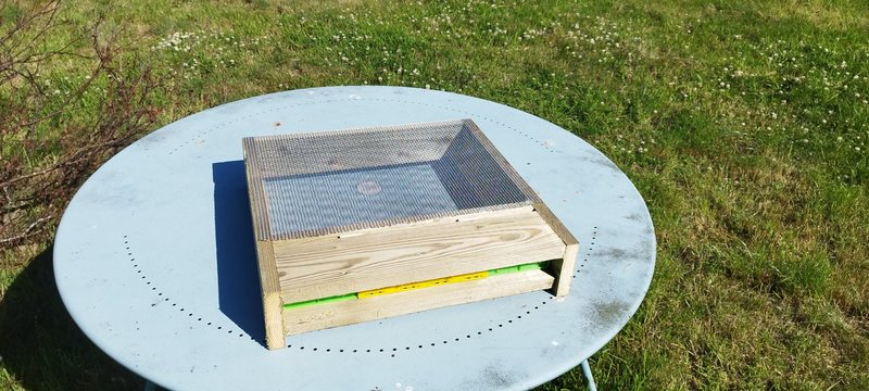 A finished timber hive floor with a mesh screen on top, sitting on a pale blue garden table on the lawn, with the green and yellow entrance slot at the front.